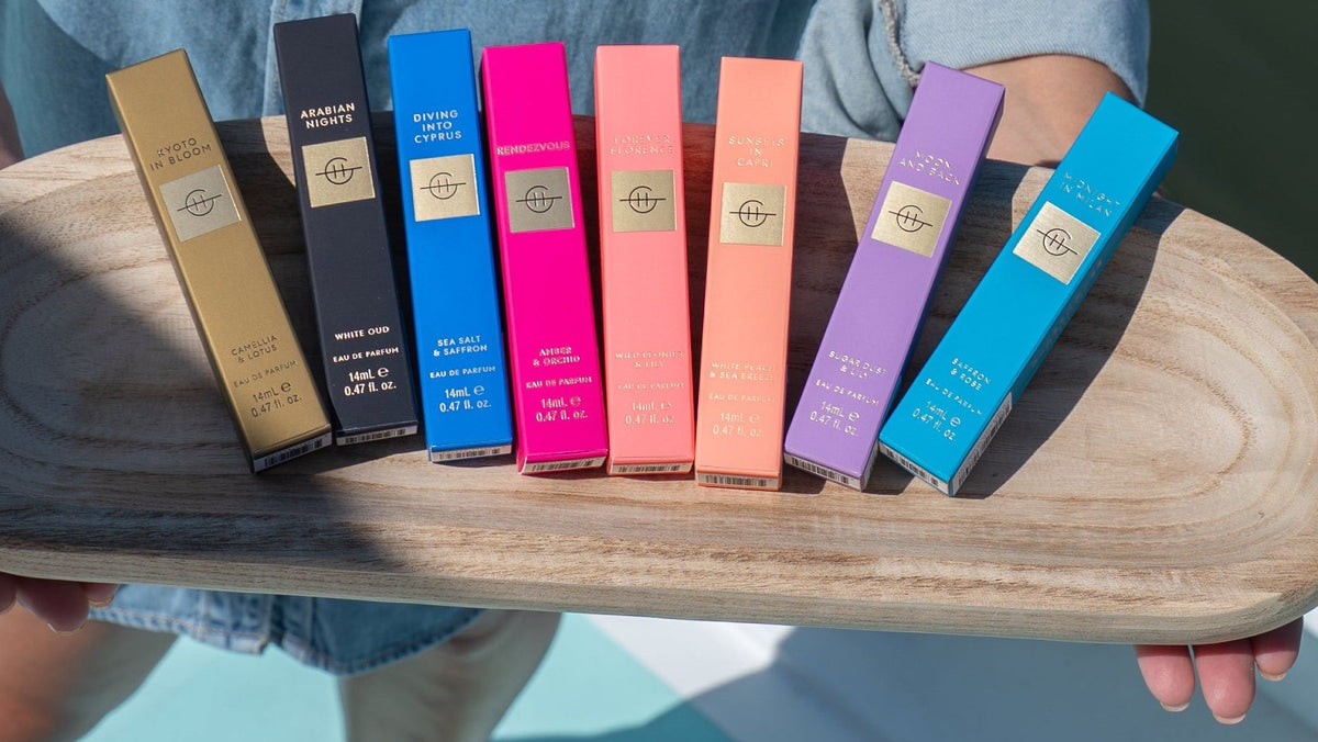 A person standing on a boat holds a wooden tray displaying seven slim rectangular boxes in different colors, including gold, navy, blue, pink, coral, purple, and light blue. The person is wearing a light blue denim shirt.