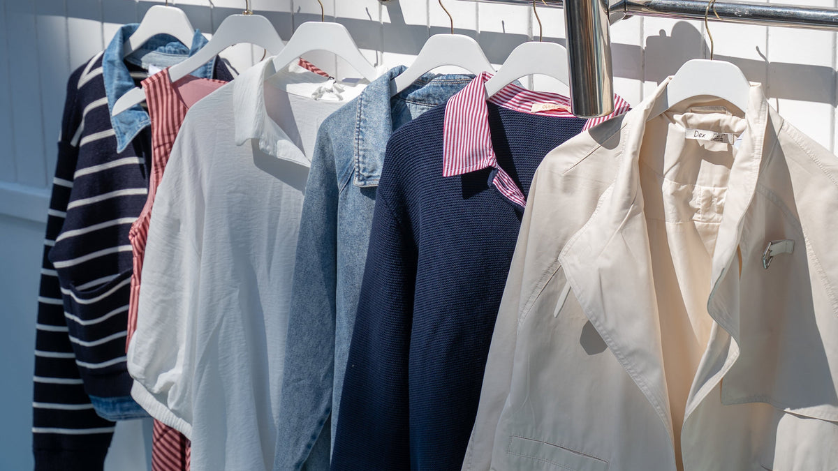 A clothing rack displaying a variety of casual garments, including a cream jacket, a navy blue jacket with a pink collar, a light blue denim shirt, a red and white striped dress, and a black and white striped sweater, set against a white paneled wall with wooden window frames.
